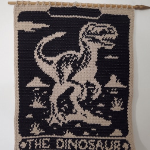 May include: Crocheted wall hanging featuring a black dinosaur silhouette against a tan background. The design includes clouds and the text "THE DINOSAUR" at the bottom. The piece is hung from a wooden dowel.
