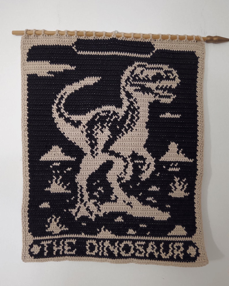 May include: Crocheted wall hanging featuring a black dinosaur silhouette against a tan background. The design includes clouds and the text "THE DINOSAUR" at the bottom. The piece is hung from a wooden dowel.
