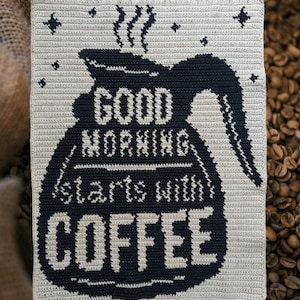 May include: A crocheted coffee-themed wall hanging. The design features a black coffee pot with steam rising, and the words "GOOD MORNING starts with COFFEE" in white lettering. The background is a light beige color, with small star accents.