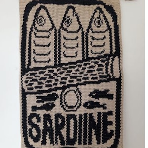 May include: A beige and black crocheted wall hanging featuring a sardine can design. The design includes three fish, a can, and the word "SARDINE" in bold letters. The wall hanging is mounted on a wooden dowel.