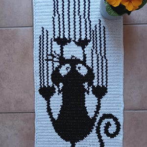 Puede incluir: Una alfombra de crochet blanca con una silueta negra de un gato. El gato se representa con una cola enroscada y se coloca en el centro de la alfombra.