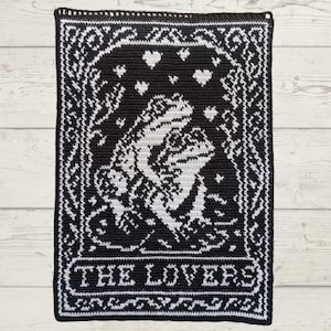 May include: A rectangular black and white crochet wall hanging featuring a graphic design of two frogs facing each other. The design includes heart shapes above the frogs and the text THE LOVERS at the bottom, all enclosed in a decorative border.