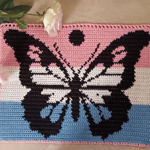 Könnte beinhalten: Ein gehäkeltes Quadrat mit einem schwarzen Schmetterlingsmuster auf einem rosa, weißen und blauen Hintergrund. Der Schmetterling befindet sich in der Mitte des Quadrats und hat ein Farbschema der Transgender-Pride-Flagge.