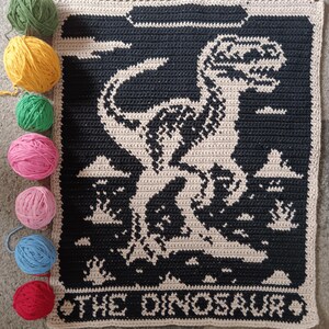 May include: A crocheted blanket featuring a black and beige dinosaur design with the text "THE DINOSAUR" at the bottom. Several balls of yarn in green, yellow, pink, blue, and red are arranged next to the blanket.