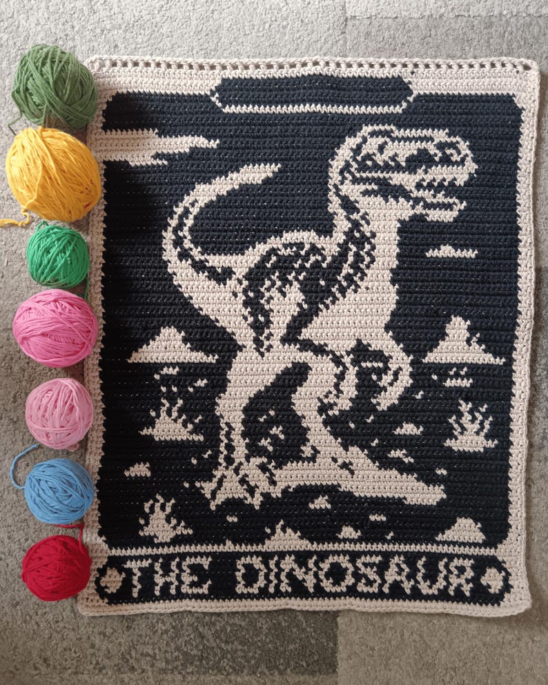 May include: A crocheted blanket featuring a black and beige dinosaur design with the text "THE DINOSAUR" at the bottom. Several balls of yarn in green, yellow, pink, blue, and red are arranged next to the blanket.