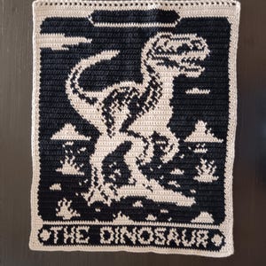 May include: A crocheted wall hanging featuring a black and white Tyrannosaurus Rex design. The dinosaur is set against a black background with a light beige border. The text "THE DINOSAUR" is at the bottom.
