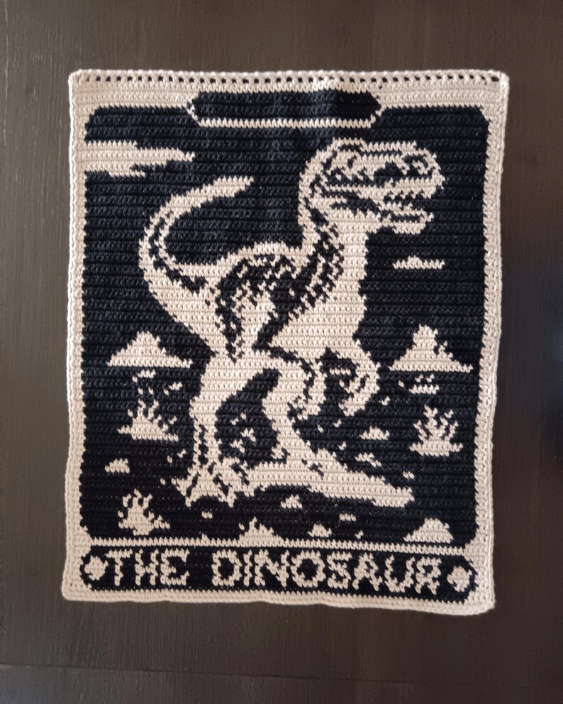 May include: A crocheted wall hanging featuring a black and white Tyrannosaurus Rex design. The dinosaur is set against a black background with a light beige border. The text "THE DINOSAUR" is at the bottom.