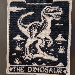 May include: A crocheted square featuring a black and cream dinosaur design. The dinosaur is centered on a black background, with the words "THE DINOSAUR" at the bottom. The edges are cream-colored.