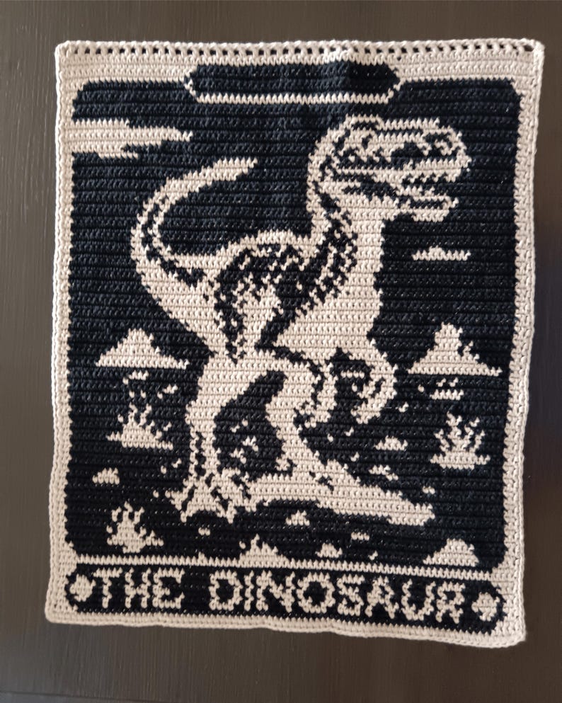 May include: A crocheted square featuring a black and cream dinosaur design. The dinosaur is centered on a black background, with the words "THE DINOSAUR" at the bottom. The edges are cream-colored.