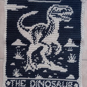 May include: Crocheted square featuring a dinosaur design in white against a navy blue background. The text "THE DINOSAUR" is at the bottom. The border is a light beige color. This handmade item is perfect for dinosaur enthusiasts.