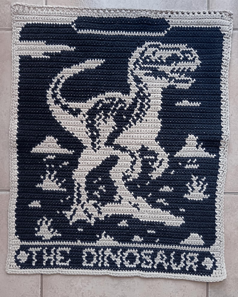 May include: Crocheted square featuring a dinosaur design in white against a navy blue background. The text "THE DINOSAUR" is at the bottom. The border is a light beige color. This handmade item is perfect for dinosaur enthusiasts.