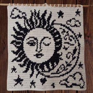 Puede incluir: Un tapiz de pared de ganchillo color crema con un diseño de sol y luna negro con estrellas y nubes. El diseño celestial está montado en una clavija de madera. El tapiz de pared se muestra sobre un fondo de madera.