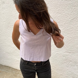 May include: A sleeveless crop top in a light purple and white gingham pattern. The top has a V-neckline and is paired with black denim jeans. The fabric appears to be lightweight, suitable for warmer weather.