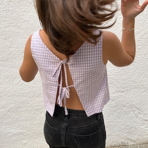 May include: A sleeveless, lilac and white gingham crop top with a low back and tie closure. The top is paired with black denim jeans. The model's hair is flowing in the wind, and a silver bracelet is visible on the wrist.