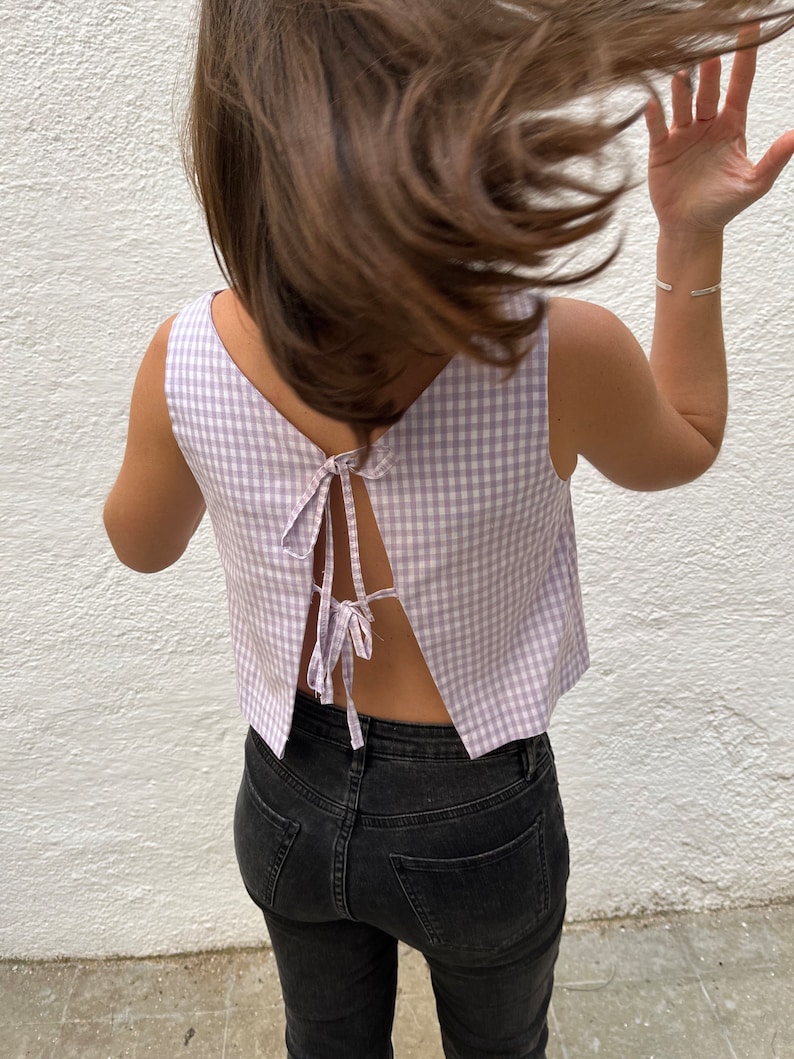 May include: A sleeveless, lilac and white gingham crop top with a low back and tie closure. The top is paired with black denim jeans. The model's hair is flowing in the wind, and a silver bracelet is visible on the wrist.