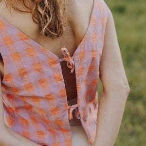 May include: A sleeveless top with a V-neck back, featuring a pink and orange checkered pattern. The top is tied at the back with small bows. The image shows the back of the top, with the person's arms and hair visible.