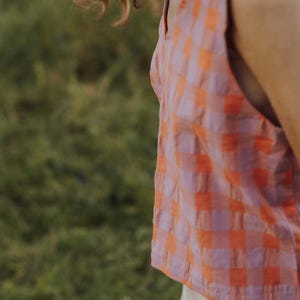May include: A sleeveless top with a pink and orange checkered pattern. The top has a scoop neckline and is paired with white pants. The background is a blurred green field.