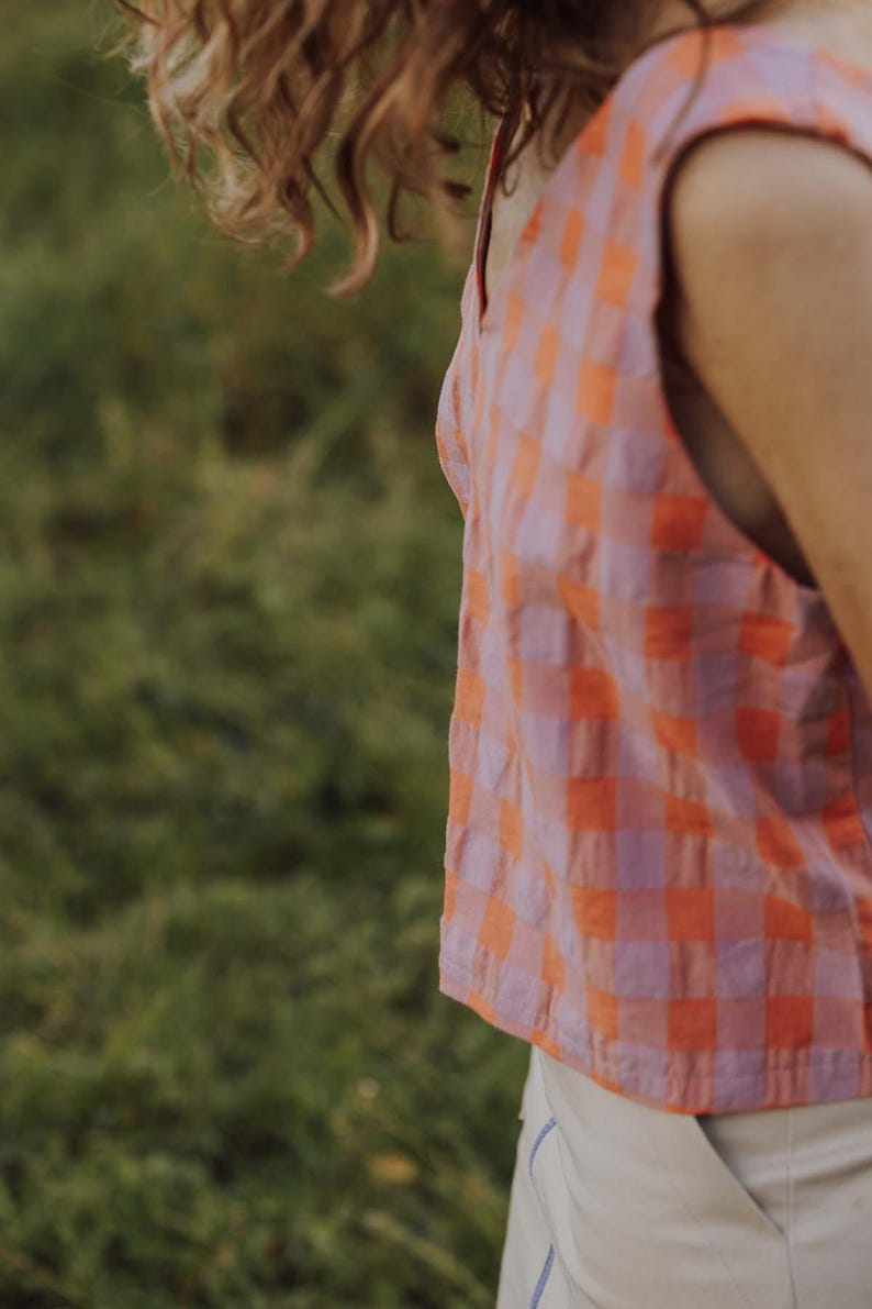 May include: A sleeveless top with a pink and orange checkered pattern. The top has a scoop neckline and is paired with white pants. The background is a blurred green field.