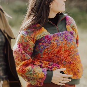 May include: A colorful fleece pullover with a half-zip closure. The jacket features a vibrant mix of orange, pink, yellow, and green hues. The sleeves and collar are a dark green color. The person is wearing a black shirt underneath and rust-colored pants.