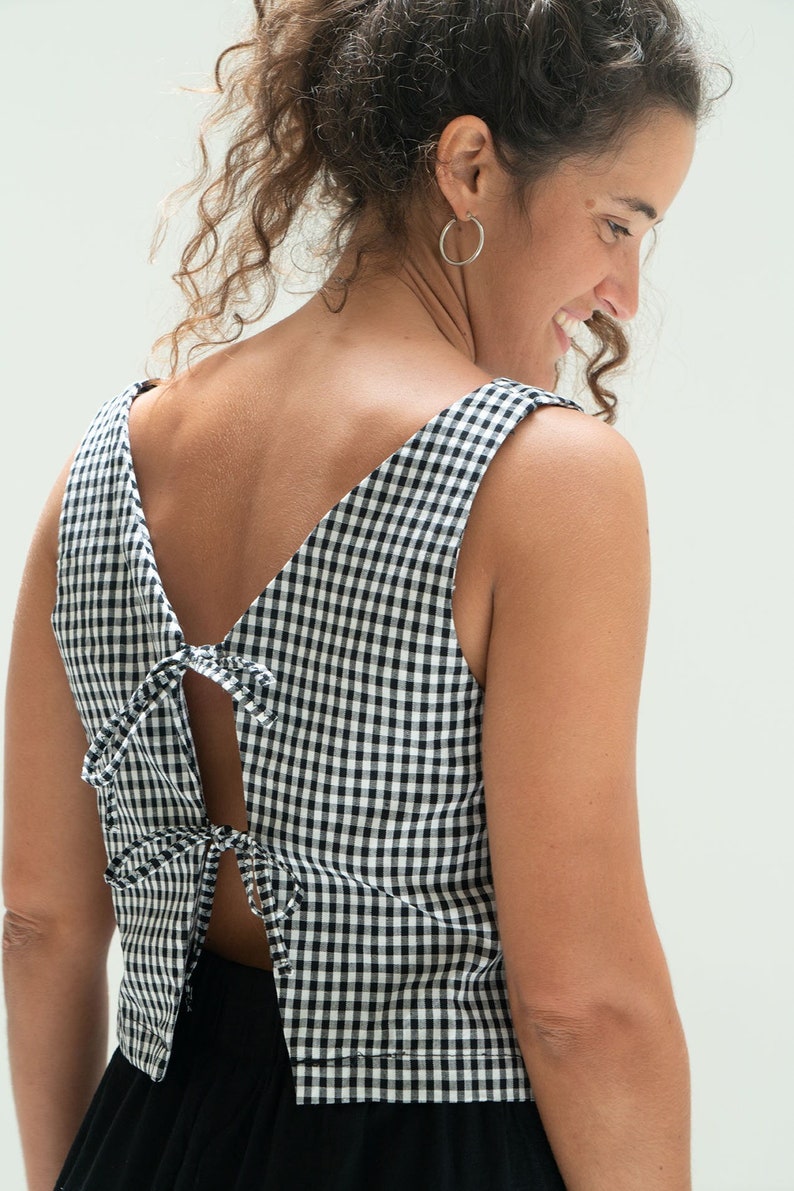 May include: A sleeveless black and white gingham top with a V-neck and open back. The back is secured with ties. The top is made of a lightweight fabric and is suitable for warm weather.