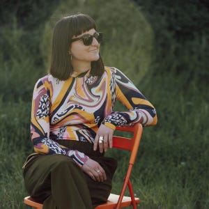May include: A woman wearing a long-sleeved top with a colorful, swirling pattern of orange, yellow, pink, and black. She is wearing sunglasses and sitting on an orange chair. The background is a grassy field.