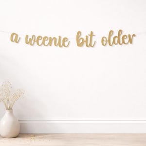 May include: A gold glittery banner featuring the cursive text a weenie bit older strung on a thin white cord. The letters are connected in a single line, creating a decorative hanging sign with a sparkling finish and a script font style.
