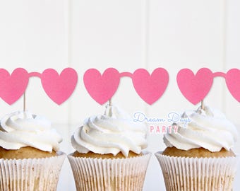 May include: Three cupcakes with white frosting, each topped with a pink heart-shaped garland. The cupcakes sit in paper liners. The text "MORE COLORS AVAILABLE" is at the bottom.