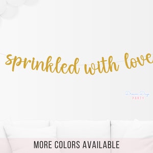 May include: Gold glitter banner with the text "sprinkled with love" hanging on a white wall. The banner is made of individual letters strung together with white string.