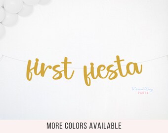 May include: Gold glitter banner with the text "first fiesta" hanging on a white wall. The banner is strung with white thread. The text "Dream Days Party" is in the bottom right corner of the image.