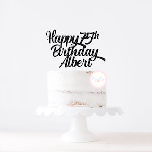 May include: A white frosted cake on a white scalloped cake stand. A black cake topper reads "Happy 75th Birthday Albert" in cursive script. A pink and blue logo is visible on the cake.