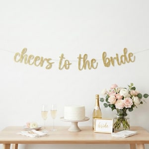 May include: A bridal shower scene with a wooden table set with a cake, champagne, flowers, and balloons. A gold glitter banner reads "Cheers to the Bride". The scene is decorated in soft, neutral colors.