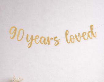 Pode incluir: Uma faixa dourada com glitter que diz "90 years loved" está pendurada em uma parede branca. Abaixo, três balões, um dourado e dois brancos. À esquerda, um vaso transparente com flores brancas.