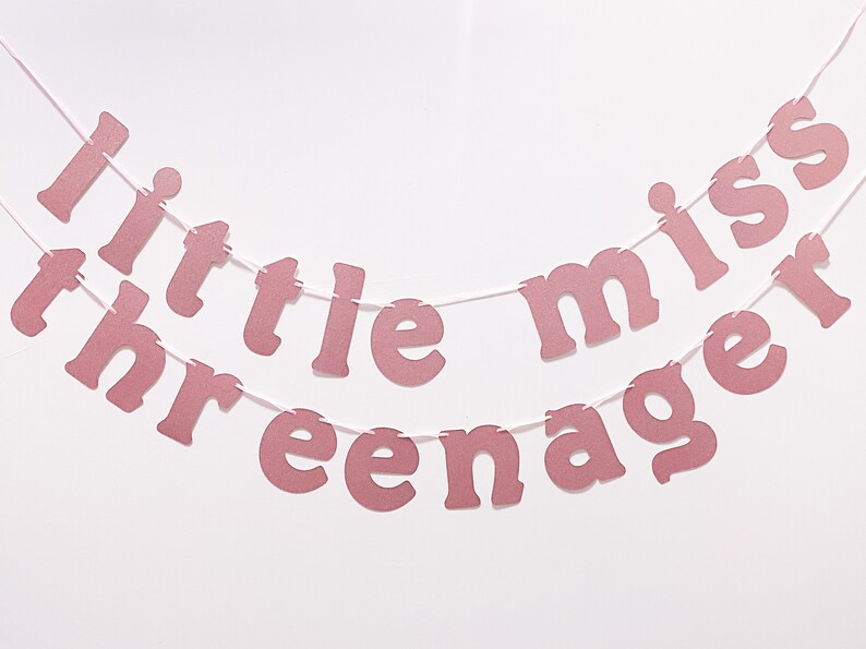 May include: Pink glitter banner with the words "little miss three" and "teenager" strung on white string. The banner is a decorative item, likely for a birthday celebration or party.