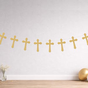 May include: A gold cross banner strung across a white wall. The banner has multiple gold crosses hanging from a white string. A vase of white flowers and three balloons are in the foreground.