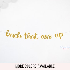May include: Gold glitter banner with the text "bach that ass up" hanging on a white wall. The banner is made of individual letters and is strung together with white string. The text is in a cursive font. The banner is a party decoration.