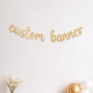 May include: A gold glitter "custom banner" strung across a white wall. The banner is in a cursive font. Below the banner are three balloons, two white and one gold. A vase of flowers is to the left. The text "LINK IN DESCRIPTION" is at the bottom.