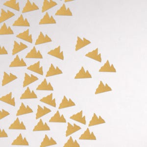 May include: A collection of small, gold-colored mountain-shaped cutouts scattered on a white background. The cutouts have a textured appearance, and the arrangement suggests a pattern or design element. The image is well-lit, with a focus on the shapes.