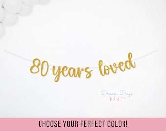 May include: A gold banner that reads "80 years loved" is strung across the image. The banner is set against a white background, with a cluster of white balloons in the upper left corner. The text "CHOOSE YOUR PERFECT COLOR!" is at the bottom.