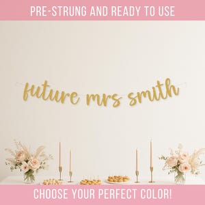 May include: A gold-colored banner featuring the text "future mrs smith" in a cursive script font, suspended on a thin string. The banner is displayed against a plain wall above a table with floral arrangements, candles, and small food items.