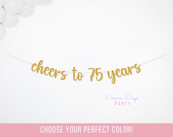 May include: A gold glitter banner that reads "cheers to 75 years" on a white string. The banner is against a white background with white balloons in the upper left corner. The text "Dream Days Party" is below the banner.