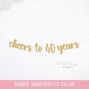 May include: A gold-colored script banner featuring the text cheers to 40 years strung on a thin white cord. The letters are cut out in a cursive font style and are suspended horizontally against a plain white background with white balloons.
