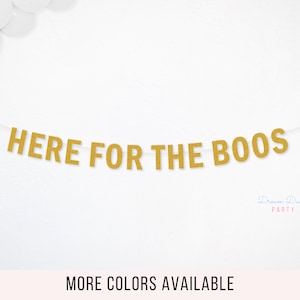 May include: A gold glitter banner featuring the text "HERE FOR THE BOOS" in capital letters, strung on a thin white cord. The letters have a textured, sparkling finish and are arranged horizontally across the center of the frame.