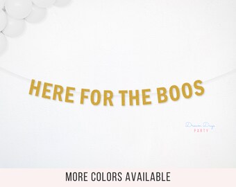 May include: A gold glitter banner featuring the text "HERE FOR THE BOOS" in capital letters, strung on a thin white cord. The letters have a textured, sparkling finish and are arranged horizontally across the center of the frame.