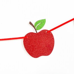 May include: A red glitter apple-shaped banner with a green glitter leaf and brown stem. The apple is strung on a red cord against a white background. This festive banner is perfect for back-to-school or fall-themed decorations.