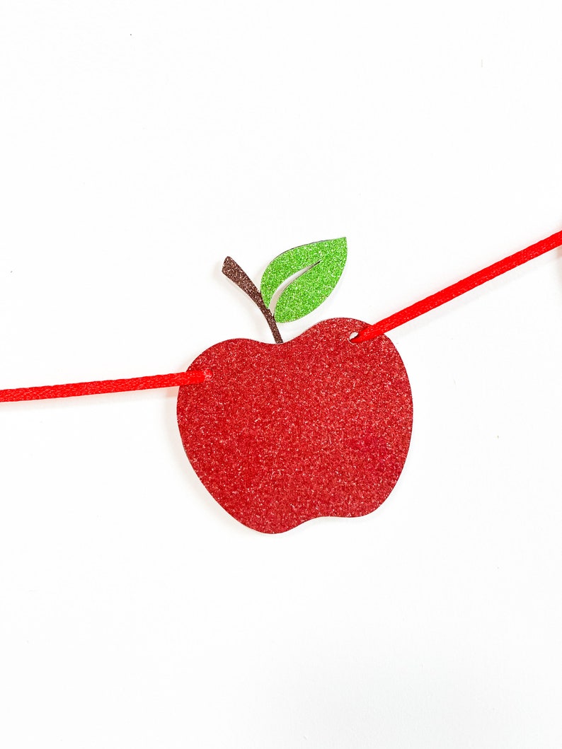 May include: A red glitter apple-shaped banner with a green glitter leaf and brown stem. The apple is strung on a red cord against a white background. This festive banner is perfect for back-to-school or fall-themed decorations.