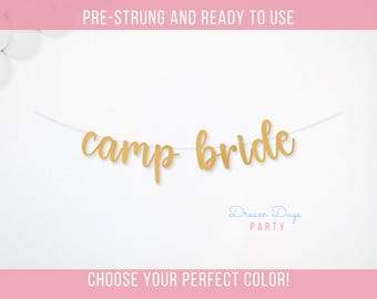 May include: A gold "camp bride" banner strung on a white string, perfect for a bridal shower or bachelorette party. The banner is set against a white background with pink accents. Text at the top reads "PRE-STRUNG AND READY TO USE".