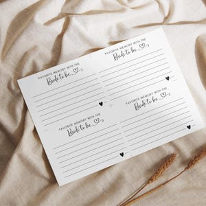 May include: A white paper card with the text "FAVORITE MEMORY WITH THE Bride to be" repeated four times, each with lined spaces for writing. Small black heart icons are scattered on the card. The card rests on a beige fabric surface, with two wheat stalks.