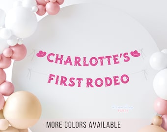 May include: A pink glitter banner that reads "CHARLOTTE'S FIRST RODEO" with cowboy hat accents. The banner is strung between two points. The background includes white, pink, and peach balloons, with the text "MORE COLORS AVAILABLE" at the bottom.