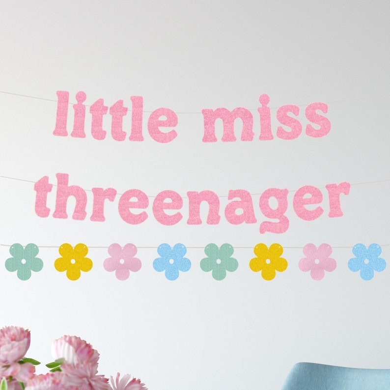 May include: A pink glitter banner with the words "little miss threenager" strung across a white background. Below the text is a garland of colorful flower cutouts in shades of blue, yellow, and pink. The banner is a festive decoration.
