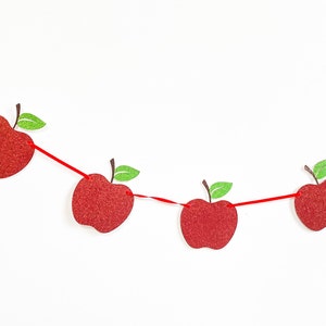 May include: A festive apple garland with red glittery apples strung on a red ribbon. Each apple has a brown stem and a green glittery leaf. The garland is set against a white background.